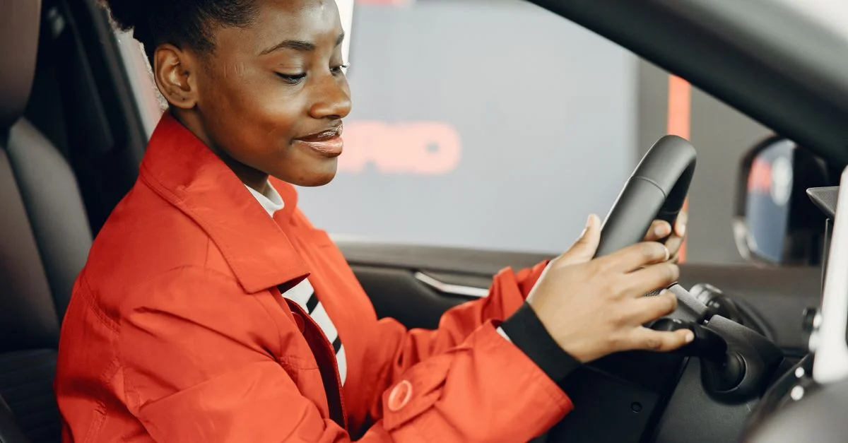 Femme noire en veste orange assise dans une voiture tenant le volant.